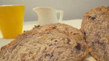 Zoom out of sliced rye raisin bread to the whole white dinner table on which there is a cup of tea, bowl of salad, white milk jug, yellow mug and some pack of honey on the front. Close-up in focus