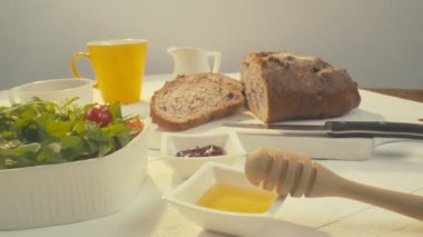 Zoom out of sliced rye raisin bread to the whole white dinner table on which there is a cup of tea, bowl of salad, white milk jug, yellow mug and some pack of honey on the front. Close-up in focus