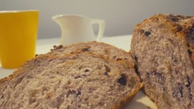 Zoom out of sliced rye raisin bread to the whole white dinner table on which there is a cup of tea, bowl of salad, white milk jug, yellow mug and some pack of honey on the front. Close-up in focus