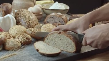 Male hands cutting the fresh baked Dutch bread on the wooden table full of other breads including rye raisin bread, wheat bread sprinkled with sesame seeds or peanuts and others