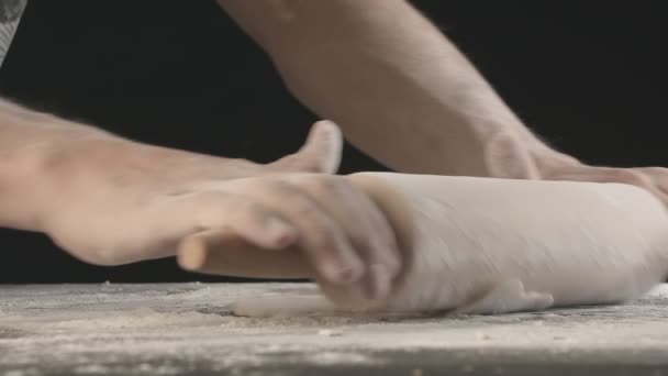 男性の手は 小麦粉を振りかけた木製のボード上の圧延ピンを使用して生地を焼くロールアウト 黒い背景で撃たれた — ストック動画