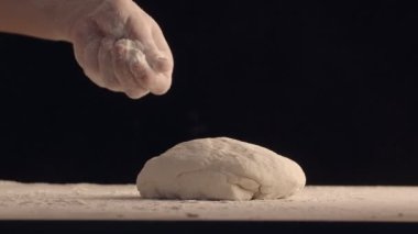 Erkek elleri bir parça hamur üzerine un serpiyor. Hamur ahşap masada yatıyor. Siyah arkaplanda çekilen.