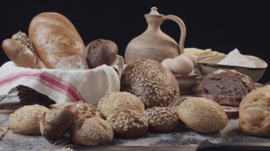 Different types of fresh baked Dutch bread is on the wooden table. wicker basket stands on the side. There are also eggs in the clay bowl, flour, butter on the back. Background is black
