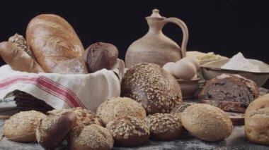 Different types of fresh baked Dutch bread is on the wooden table. wicker basket stands on the side. There are also eggs in the clay bowl, flour, butter on the back. Background is black