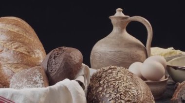Different types of fresh baked Dutch bread is on the wooden table. wicker basket stands on the side. There are also eggs in the clay bowl, flour, butter on the back. Background is black