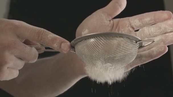 小さな金属ふるいを通して小麦粉をふるいにかける男性の手のクローズアップ 黒を背景に撮影 — ストック動画