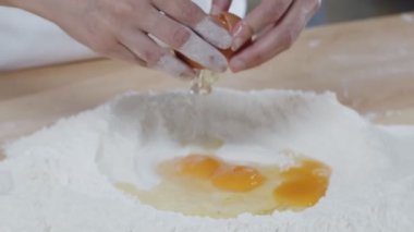 Baker break eggs to the flour lying on the wooden cooking table. Slow motion