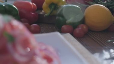 Close-up camera moves from fresh veggies and greens including red, green and yellow bell peppers, radish and juniper on the wooden cooking board to the salad made of tomatoes and red onions