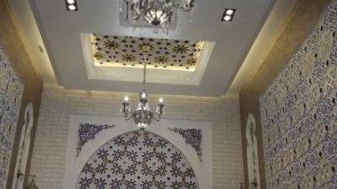 Niche in an oriental restaurant with white walls ornate with gold and blue floral ornaments. There is a white long table set for dinner in the room. Stools are also white with purple seats
