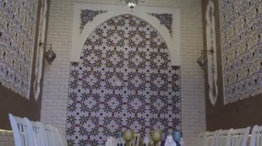 Niche in an oriental restaurant with white walls ornate with gold and blue floral ornaments. There is a white long table set for dinner in the room. Stools are also white with purple seats
