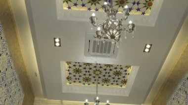 Niche in an oriental restaurant with white walls ornate with gold and blue floral ornaments. There is a white long table set for dinner in the room. Stools are also white with purple seats