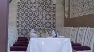 Niche in an oriental restaurant with white walls ornate with gold and blue floral ornaments. There is a white long table set for dinner in the room. Stools are also white with purple seats
