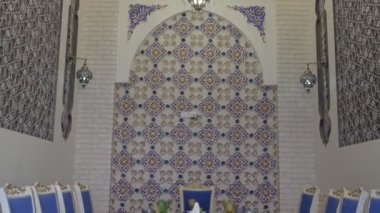 Niche in an oriental restaurant with white walls ornate with gold and blue floral ornaments. There is a white long table set for dinner in the room. Stools are also white with purple seats