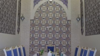 Niche in an oriental restaurant with white walls ornate with gold and blue floral ornaments. There is a white long table set for dinner in the room. Stools are also white with purple seats