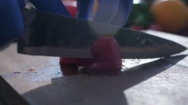 Close-up of tomato cut by the knife on the wooden cooking board. The hands of a cook are in blue rubber gloves