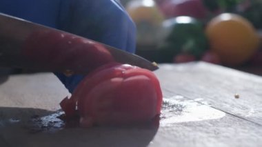 Close-up of tomato cut by the knife on the wooden cooking board. The hands of a cook are in blue rubber gloves