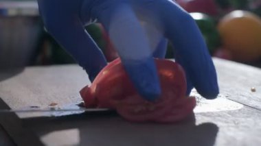 Close-up of tomato cut by the knife on the wooden cooking board. The hands of a cook are in blue rubber gloves