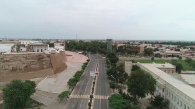 Street near the front gate Ark of old Bukhara city fortress a filmed by drone on a warm cloudy day. Mild traffic on the road and waking people