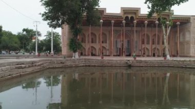 And old eastern building with high wooden carved columns on the pediment placed near the artificial pond. Filmed in old Bukhara city on cloudy day.