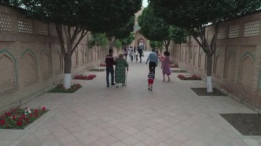 Enterance of memorial complex of Bahouddin Naqshband near Bukhara. Drone cam films the small cobbled walled alley with trees on the sides where visitors walk then it rises up and we see gravestones