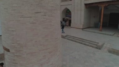 Drone cam is rising near the small minaret column in the middle of the yard of memorial complex of Bahouddin Naqshband near Bukhara. At-first we see several passers-by and the door of a gift shop