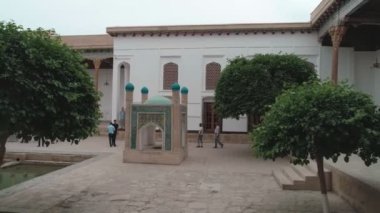 Drone cam films the inner-yard of memorial complex of Bahouddin Naqshband near Bukhara. There are decorations, trees, pool and benches where visitors sit. Carved columns are in front of the camera