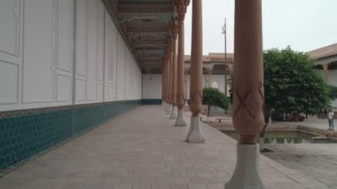 Drone cam films the inner-yard of memorial complex of Bahouddin Naqshband near Bukhara. There are decorations, trees, pool and benches where visitors sit. Carved columns are in front of the camera