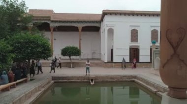 Drone cam films the inner-yard of memorial complex of Bahouddin Naqshband near Bukhara. There are decorations, trees, pool and benches where visitors sit. Carved columns are in front of the camera