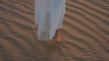 A young woman in a white dress barefoot is walking along the desert sand. Slow motion.