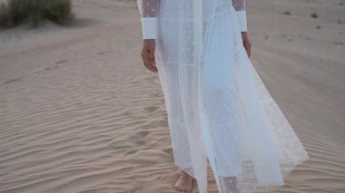 A young woman in a white dress barefoot is walking along the desert sand. Slow motion.