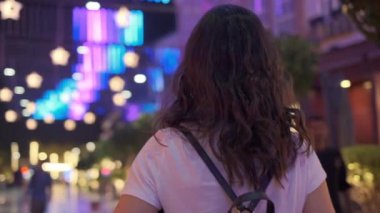 A young beautiful woman walks alone along a brightly lit night street.