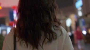 A young beautiful woman walks alone along a brightly lit night street.