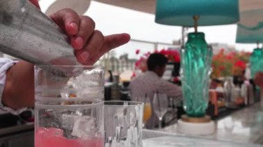 Barman puts ice into coctail shaker. He is wearing white summer shirt with doodles. Slow motion.