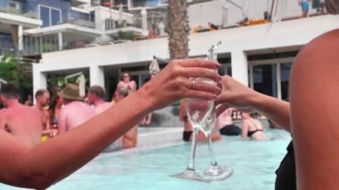 Two female hands clink glasses against the background of the pool. There is water in glasses, splashes of which fly to the sides.