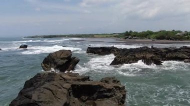 Waves rush Balis rocky Overgrown Cliffs. Weather is windy. There are some ancient walls and buildings near the coastal line.