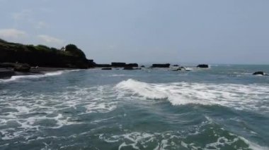 Waves rush Balis rocky Overgrown Cliffs. Weather is windy. There are some ancient walls and buildings near the coastal line.