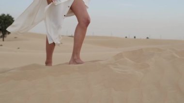A brunette barefoot in a white dress fluttering in the wind walks along the desert sand. Slow motion.