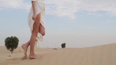 A brunette barefoot in a white dress fluttering in the wind walks along the desert sand. Slow motion.