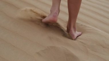 A brunette barefoot in a white dress fluttering in the wind walks along the desert sand. Slow motion.