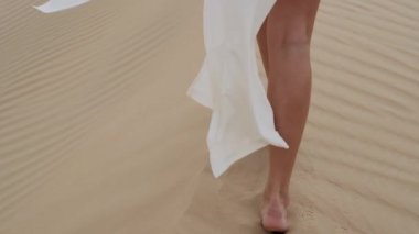 A brunette barefoot in a white dress fluttering in the wind walks along the desert sand. Slow motion.