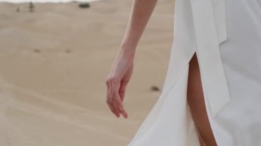 A brunette barefoot in a white dress fluttering in the wind walks along the desert sand. Slow motion.