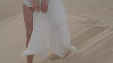 A brunette barefoot in a white dress fluttering in the wind walks along the desert sand. Slow motion.