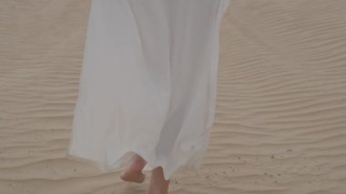 A brunette barefoot in a white dress fluttering in the wind walks along the desert sand. Slow motion.