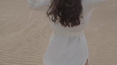 A brunette barefoot in a white dress fluttering in the wind walks along the desert sand. Slow motion.
