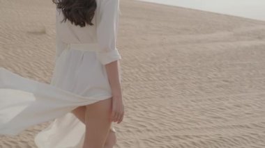 A brunette barefoot in a white dress fluttering in the wind walks along the desert sand. Slow motion.
