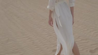 A brunette barefoot in a white dress fluttering in the wind walks along the desert sand. Slow motion.