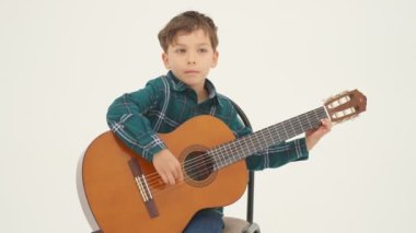 Küçük çocuk taburede oturmuş büyük klasik gitar çalıyor. Yavaş çekim