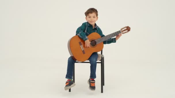 小さな男の子の大きな古典的なギターのスツールに座って演奏されます スローモーション — ストック動画