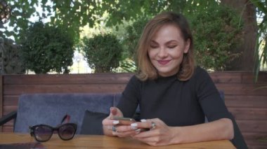 Woman wearing black sits by the table and looks at smartphone. Filmed outdoors