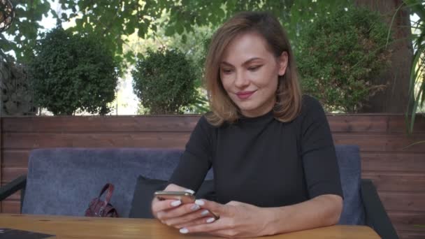 Woman Wearing Black Sits Table Looks Smartphone Filmed Outdoors — Vídeos de Stock
